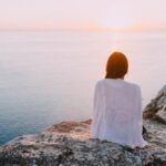 donna seduta su una scogliera che guarda il tramonto sul mare